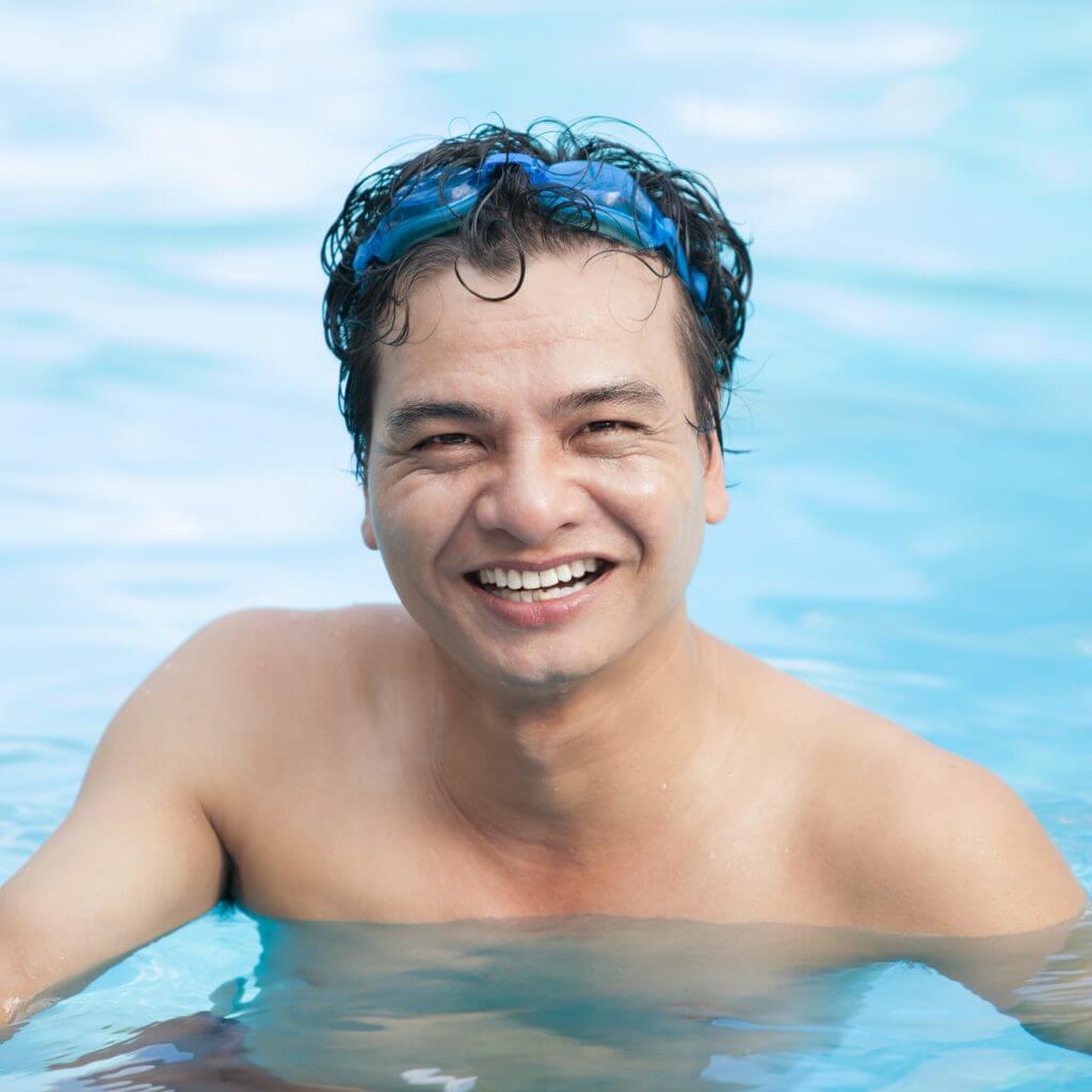 middle aged Asian man in pool with swim goggles on his head, smiling at the camera.