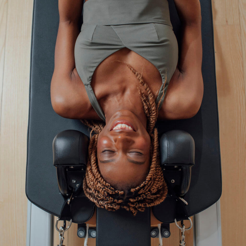 From above photo of pretty African smiling woman in sportswear lying on pilates reformer.