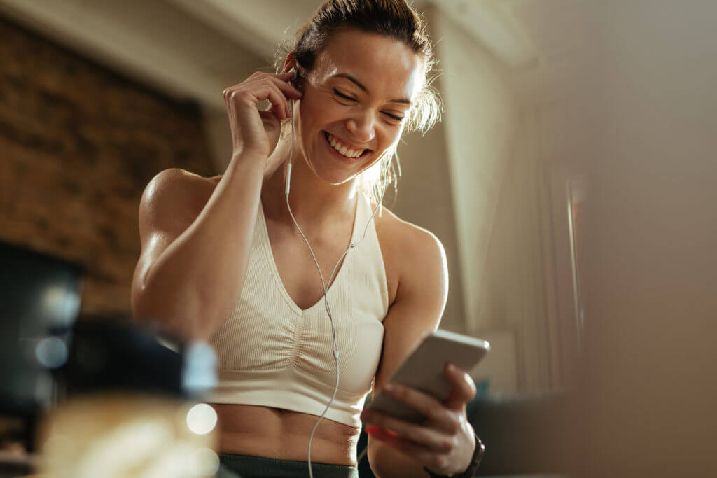 Young happy sportswoman using mobile phone while listening music over earphones.