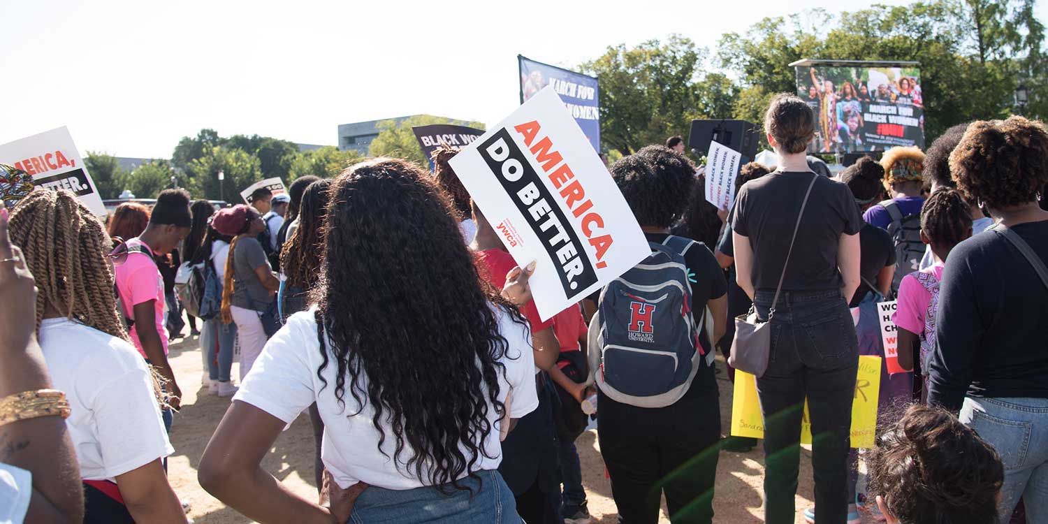 from behind, looking at a crowd of people at a rally (a podium is visible in the far background). In the center of the frame is a Black woman with long, dark hair holding a sign that says "America, do better."