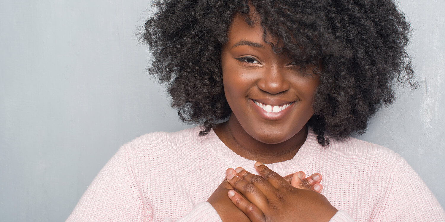 Young african american plus size woman over grey grunge wall wearing winter sweater smiling with hands on chest with closed eyes and grateful gesture on face.