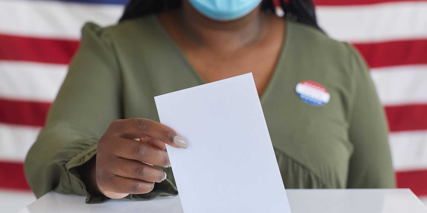 Close up of African-American woman wearing mask putting vote bulletin in ballot box and looking at camera while standing against American flag on election day
