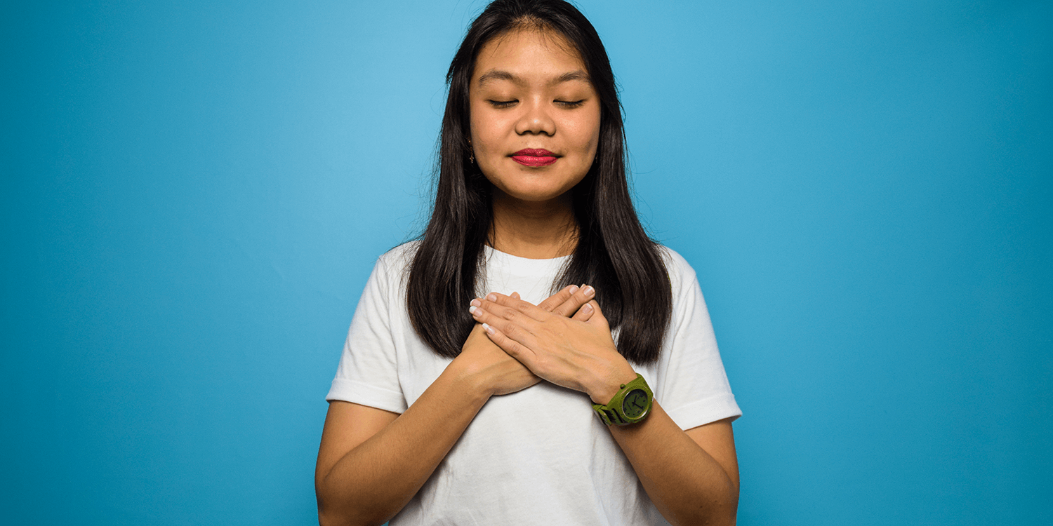 Portrait of Young Asian women using white T-shirt with blue isolated background, smiling with hands on chest with closed eyes and grateful