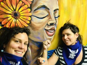 Artists Camila Leiva and Claudia Valentino smiling together in front of the mural they created titled Loving Yourself is Revolutionary at a YWCA St. Paul housing program facility